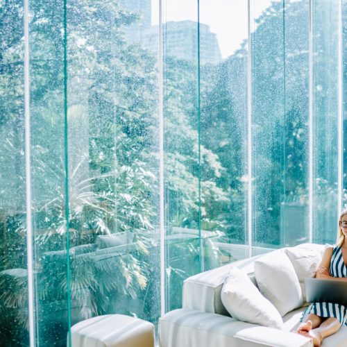 Happy female freelancer with red lips in glasses looking at camera and using laptop while resting at comfortable sofa in contemporary workspace with glass walls