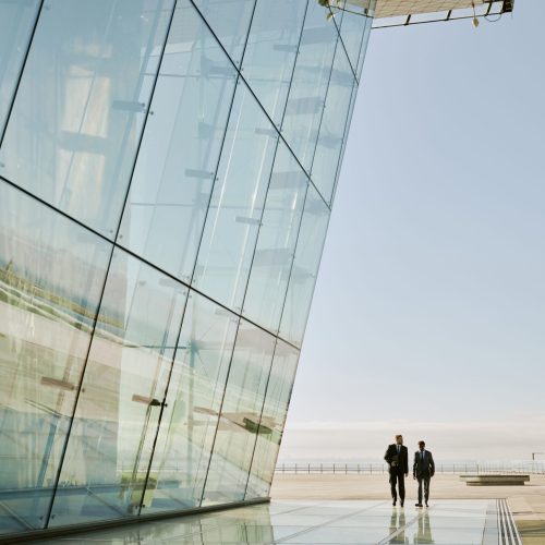Reflective glass facade of modern building casting elegant patterns onto the surrounding urban landscape showing two individuals walking along the mirrored surface in sunlight